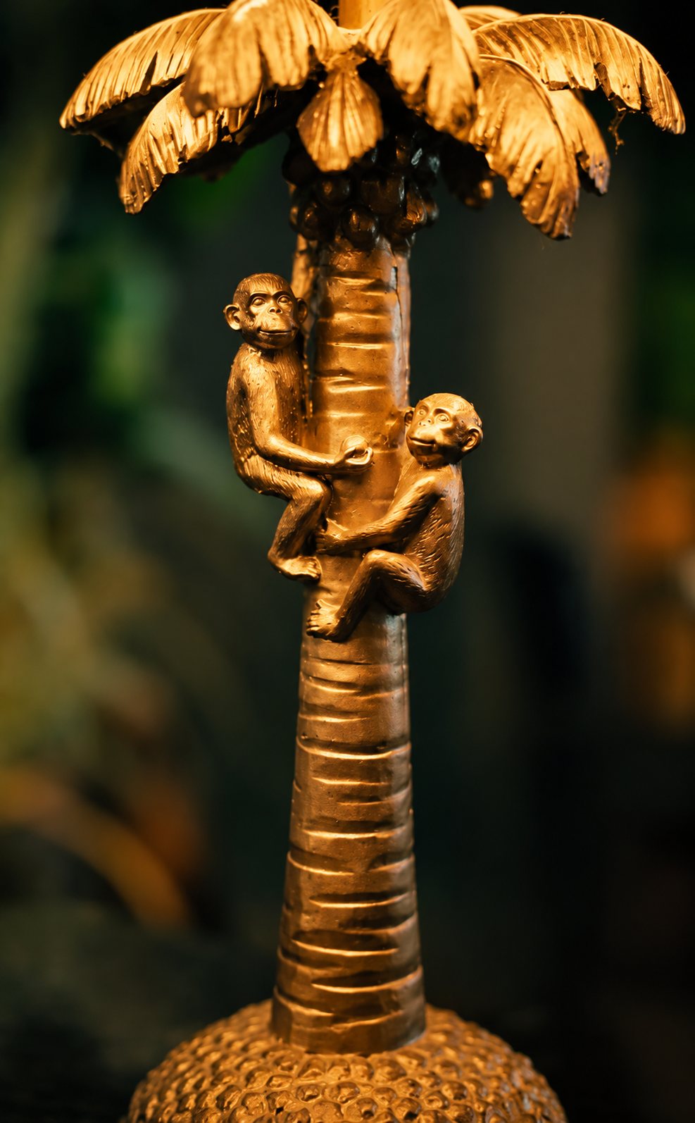 Gros plan sur un objet décoratif doré en forme de palmier sur lequel grimpent deux petits singes, devant un arrière-plan sombre et flou.