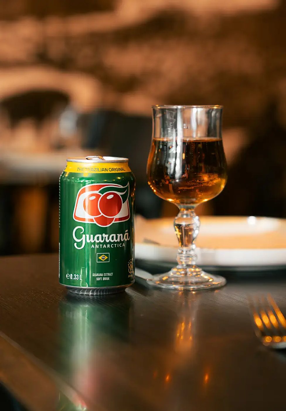 Une canette verte de Guaraná Antarctica posée sur une table en bois sombre, à côté d'un verre à pied rempli de boisson ambrée, dans un restaurant chaleureux.