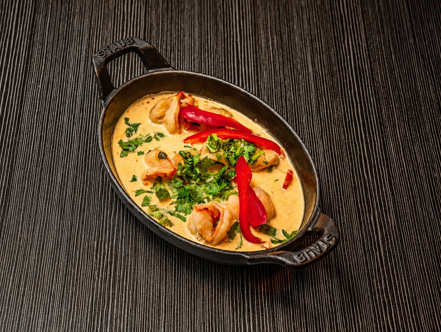 Une petite cocotte ovale en fonte noire contient des crevettes dans une sauce crémeuse au curry, garnie de coriandre fraîche et de lanières de poivron rouge, posée sur une table en bois sombre.