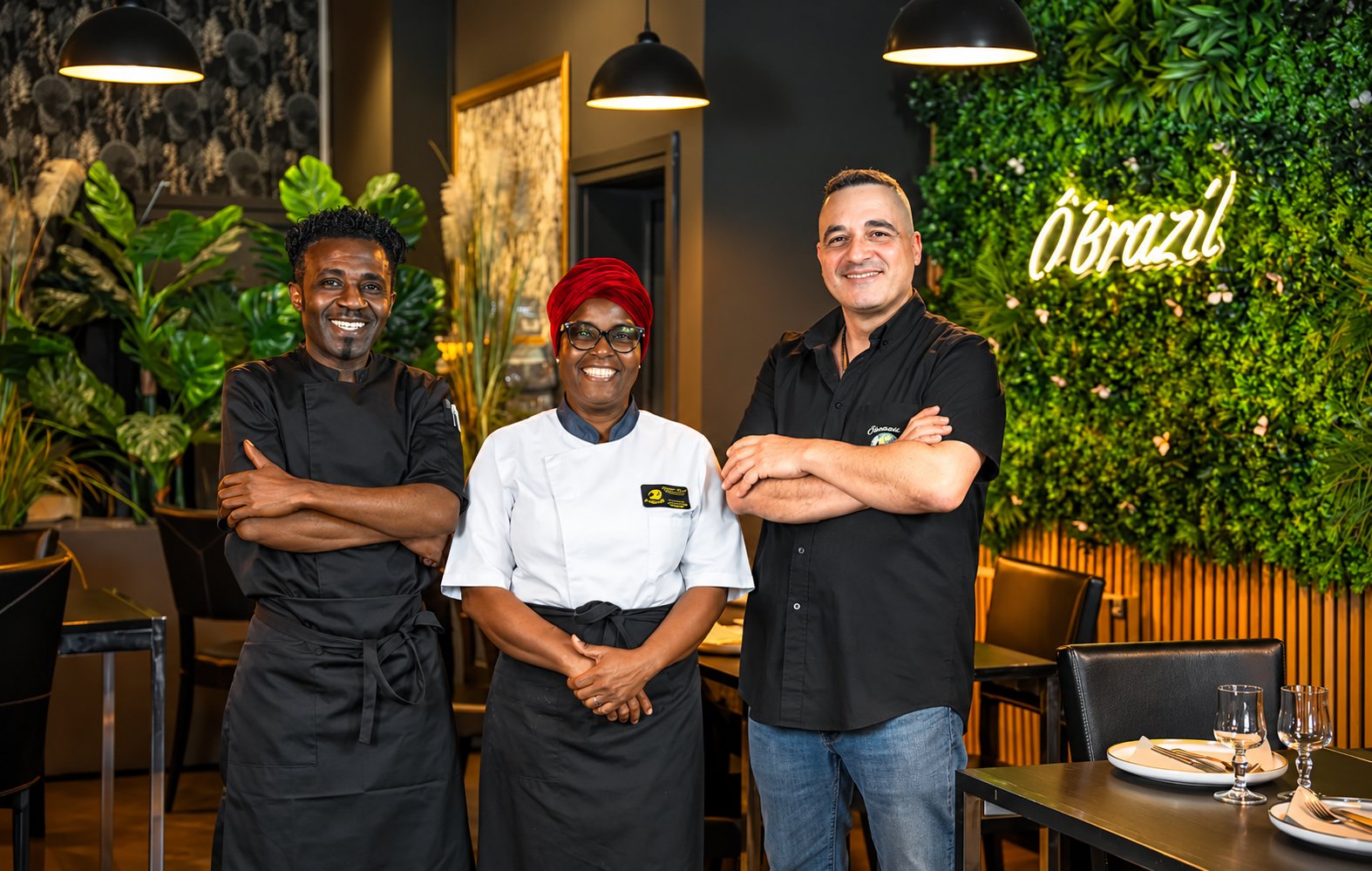 Trois membres de l'équipe du restaurant, deux en tenue de cuisine et un en chemise noire, posent les bras croisés devant un mur végétal orné d'une enseigne néon.