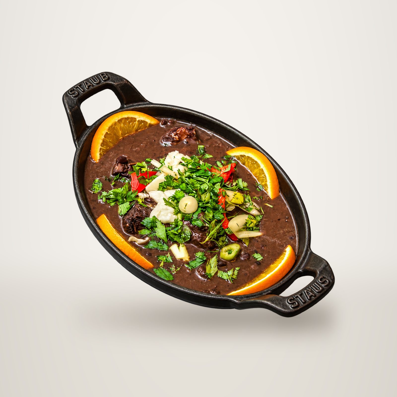 Un plat ovale en fonte noire contient une feijoada de haricots noirs garnie d'herbes fraîches, de piment rouge et de tranches d'orange.
