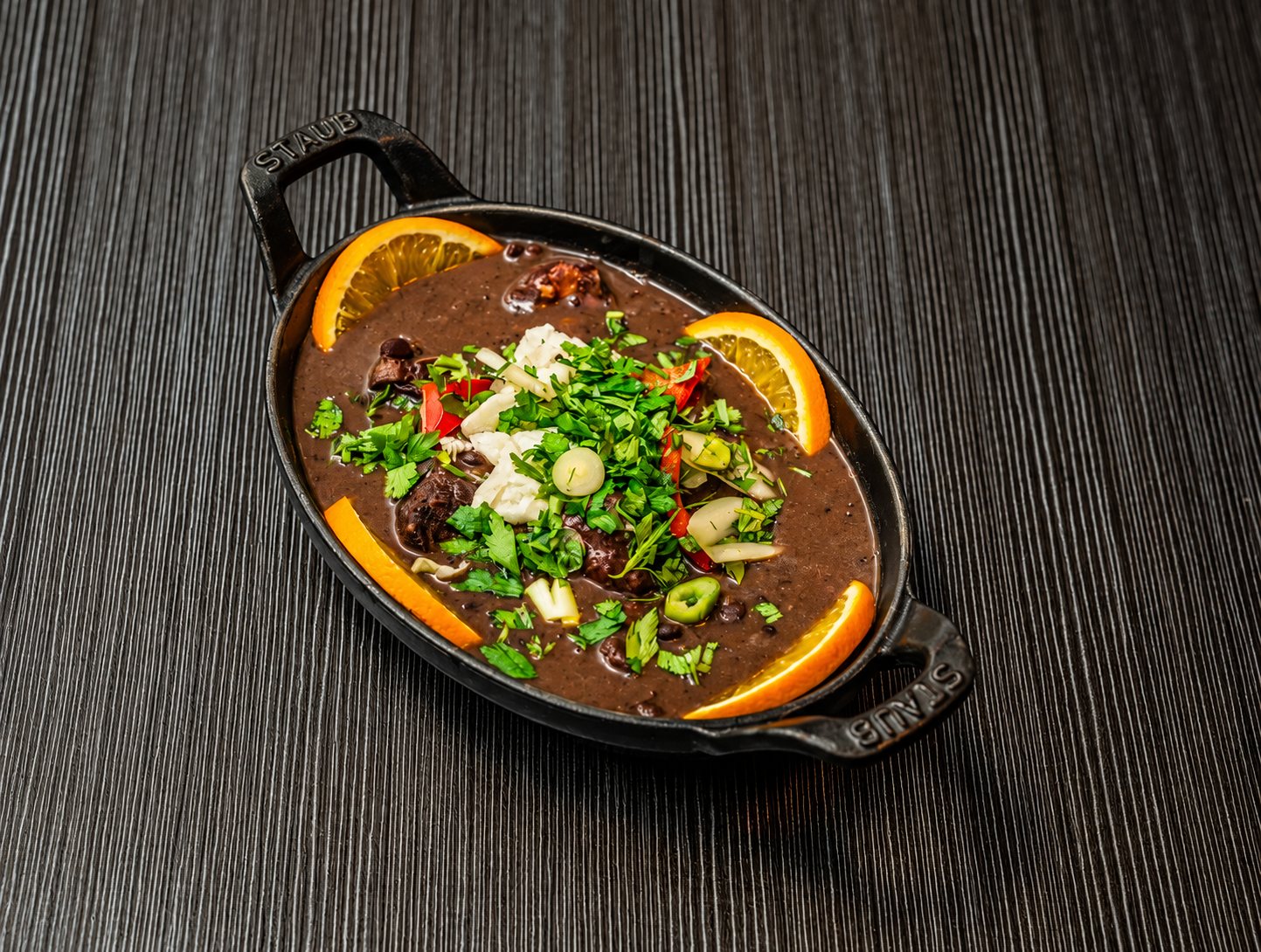 Cocotte ovale en fonte noire remplie d'un ragoût de haricots noirs garni de coriandre, de piment rouge, de fromage frais et de tranches d'orange, posée sur une table en bois sombre.