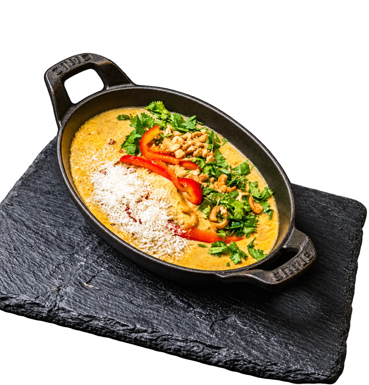 Une poêle en fonte noire contient un plat crémeux au curry garni de poivron rouge, d'herbes fraîches, de cacahuètes et de fromage râpé, posée sur une ardoise.