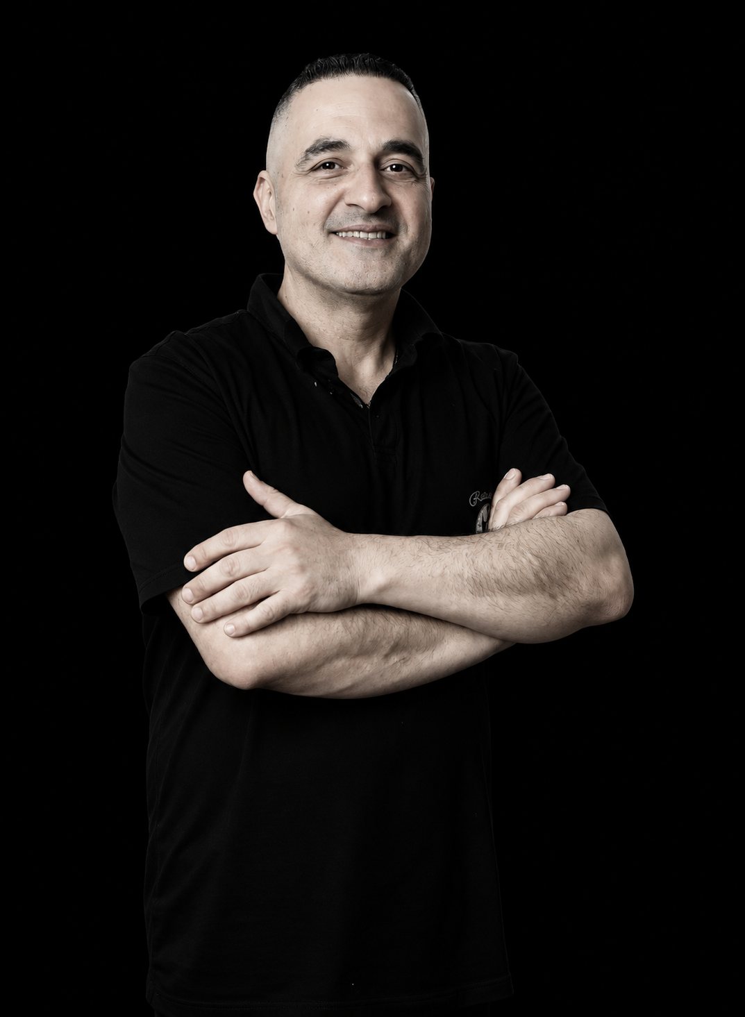Portrait en noir et blanc d'un homme souriant en polo sombre, les bras croisés, sur fond noir.