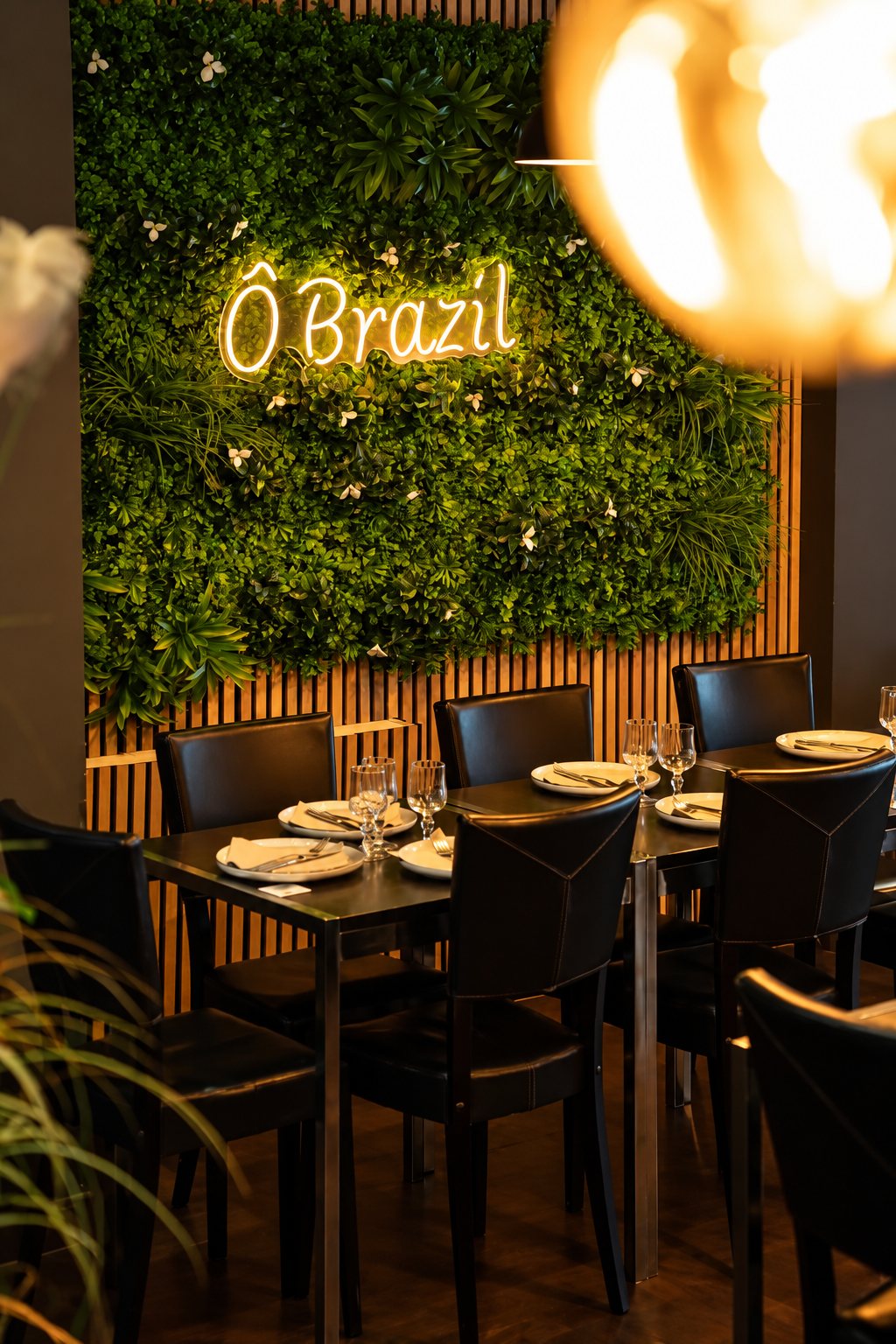 Tables dressées avec assiettes et verres devant un mur végétal éclairé par l'enseigne néon lumineuse Ô Brazil.