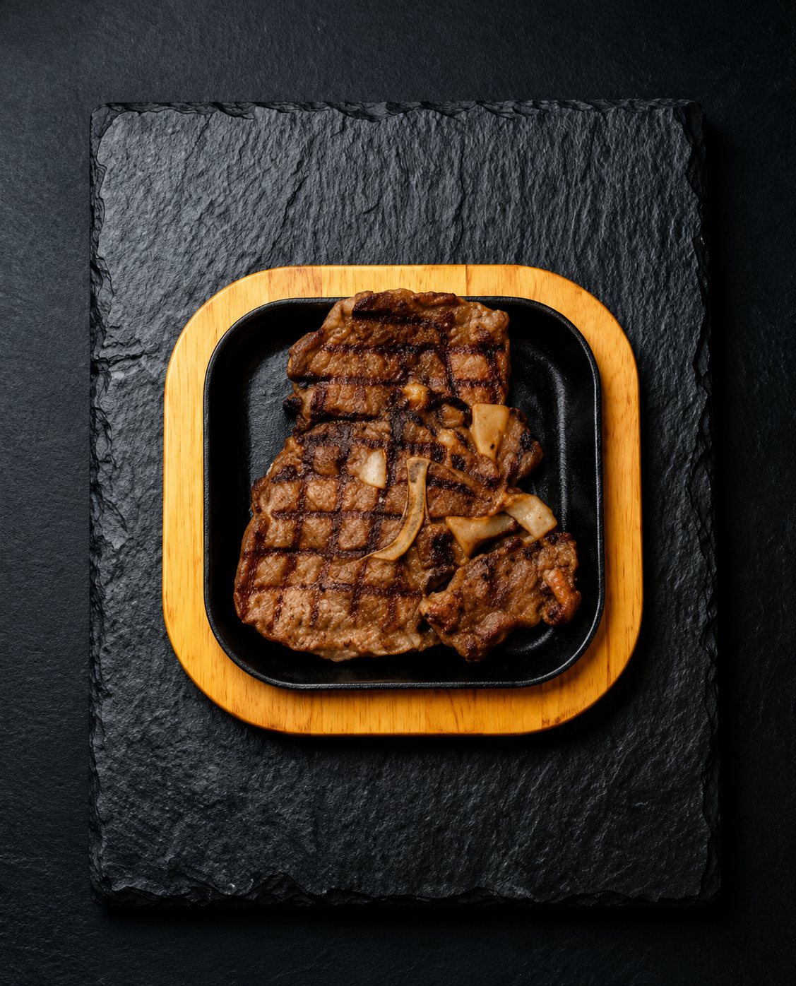 Vue de dessus d'un steak grillé aux oignons posé sur un plateau en bois carré, lui-même placé sur une ardoise noire.