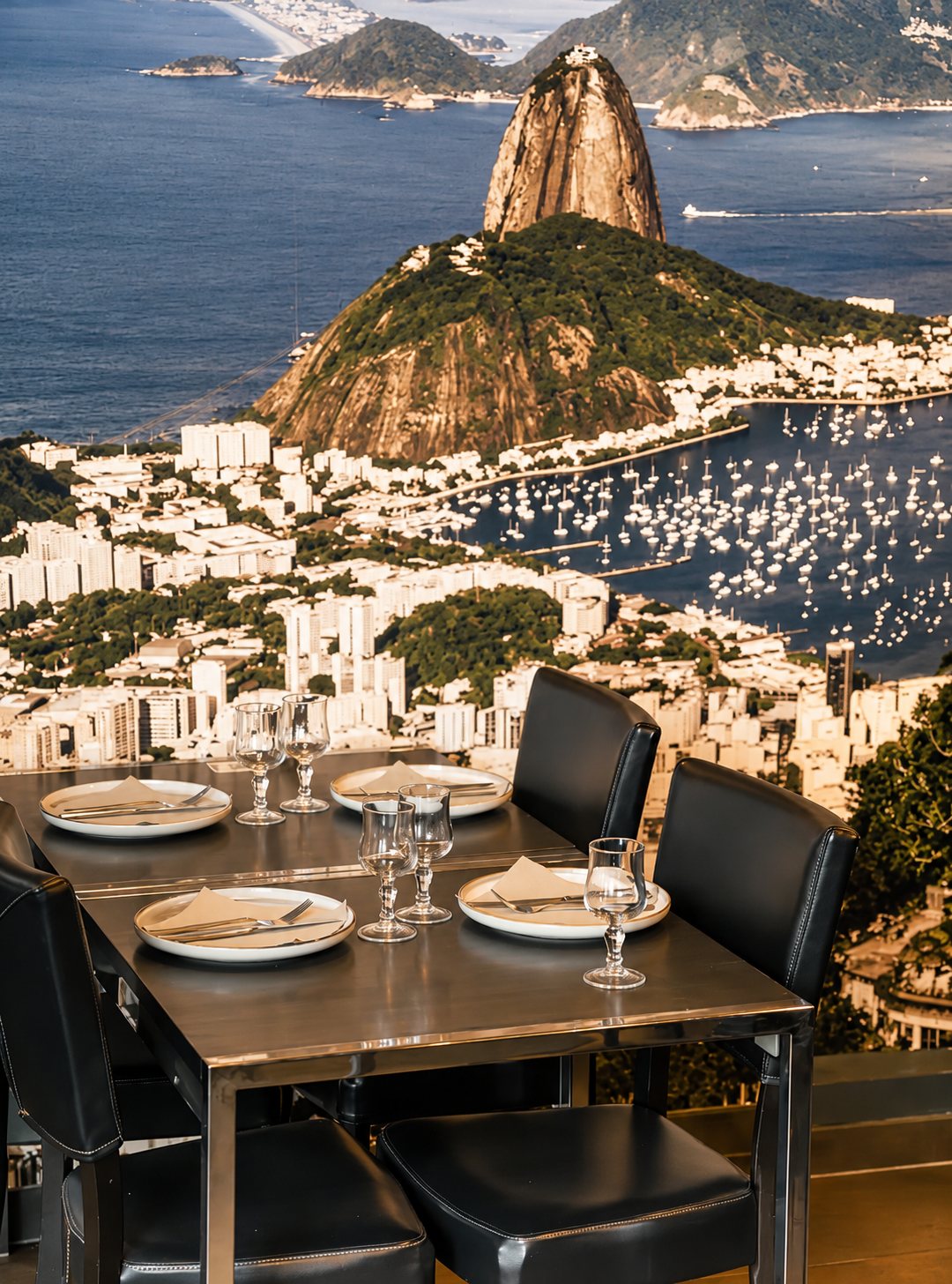 Une table de restaurant dressée avec assiettes, verres et chaises en cuir noir devant une grande photo murale en tons sépia d'une baie avec le Pain de Sucre.